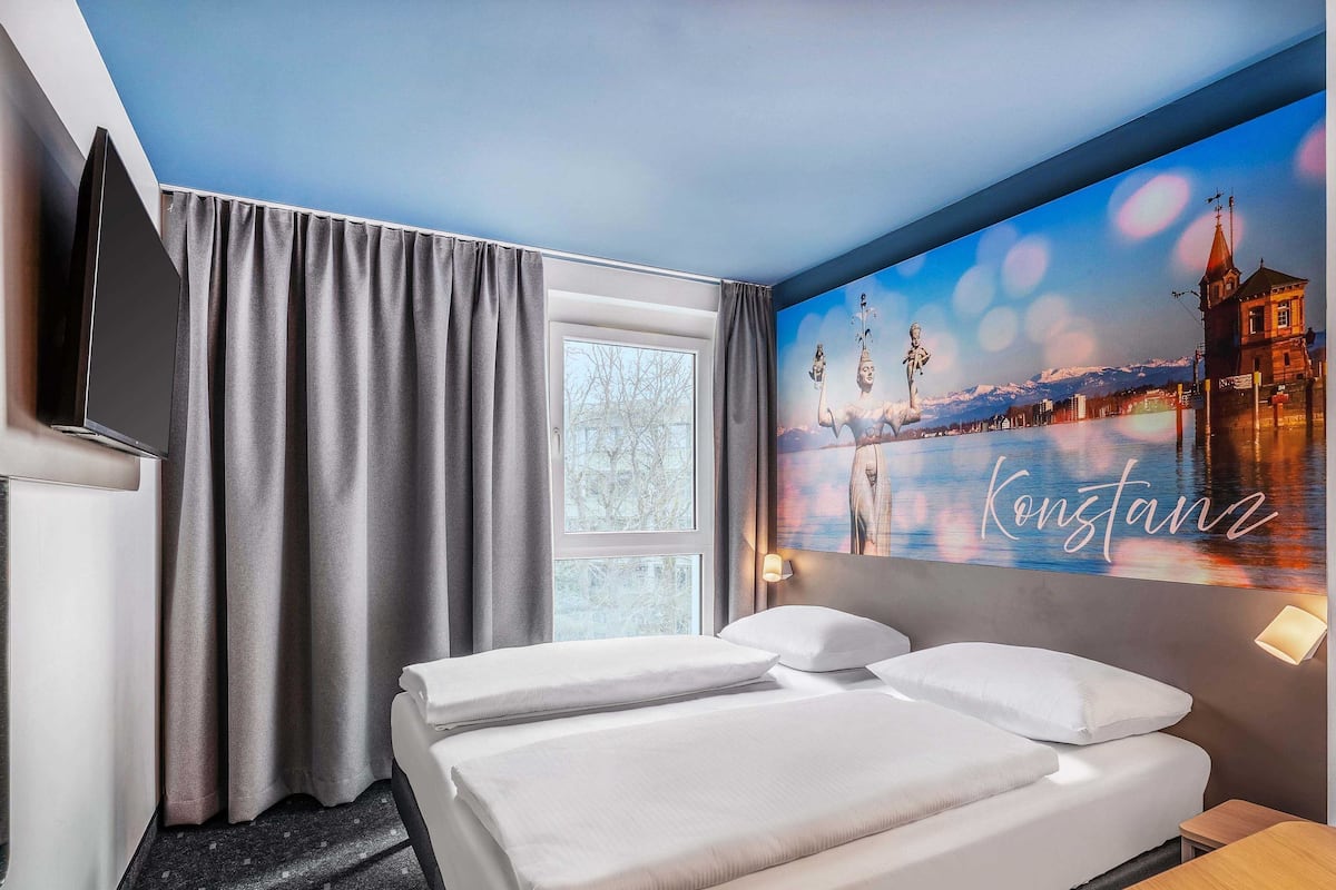 A hotel room with a bed, a TV, a window with curtains, and a wall with a cityscape painting.