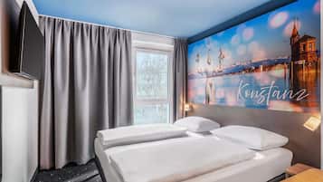 A hotel room with a bed, a TV, a window with curtains, and a wall with a cityscape painting.