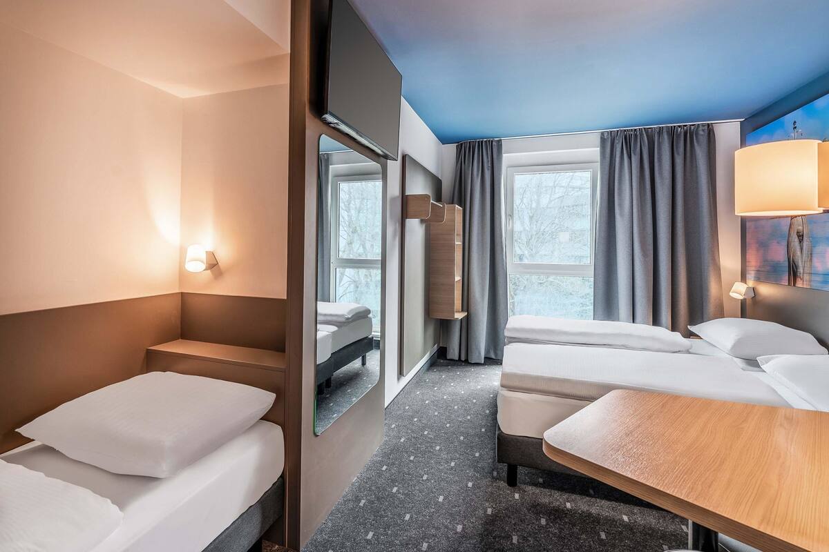 A hotel room with two beds, a mirror, a window with curtains, a lamp, and a small wooden table.
