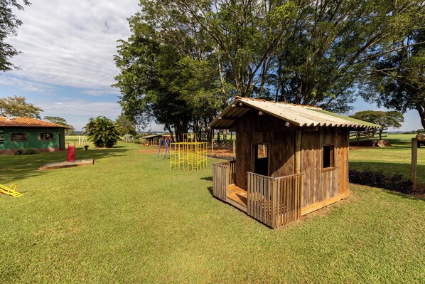 A small wooden playhouse with a porch, surrounded by a grassy area with trees and a clear sky.