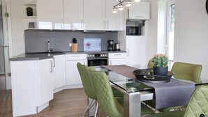 A modern kitchen with white cabinets, a built-in oven, and a dining area with green chairs and a glass table. - Haus Nina - Sonnenweg 20 (Diemelsee)