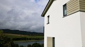 A modern building with a white exterior and beige siding, overlooking a lake and hilly landscape. - Haus Nina - Sonnenweg 20 (Diemelsee)