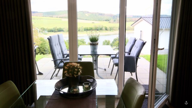 A dining area with a table, chairs, and a view of a lake and greenery. - Haus Nina - Sonnenweg 20 (Diemelsee)