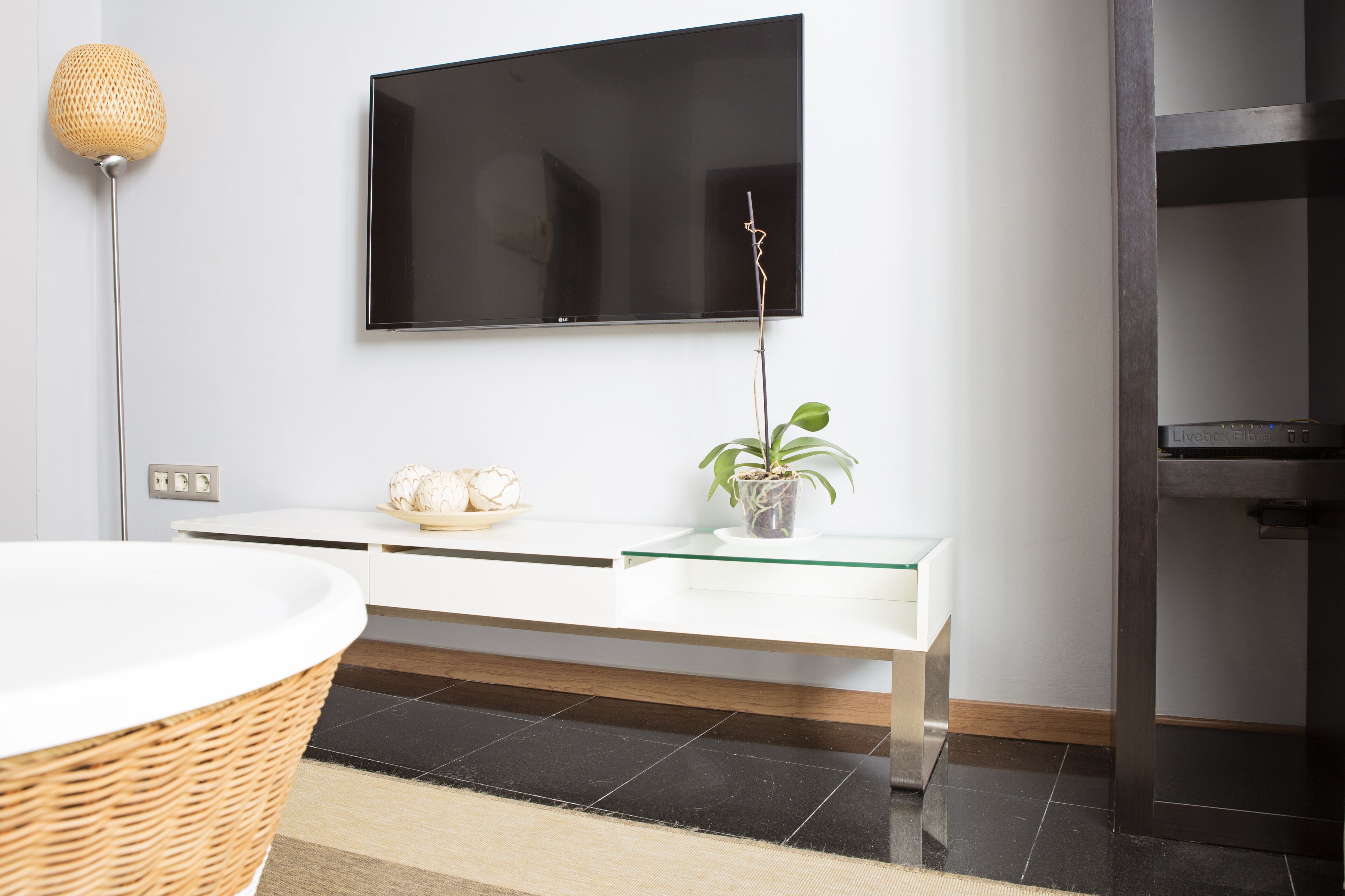 A modern bathroom with a large mirror, a white vanity, a flat-screen TV mounted on the wall, and a potted plant on the vanity.