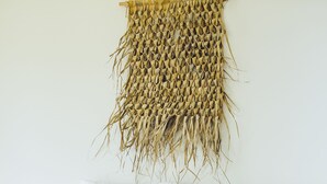 Un lit recouvert de draps blancs, une tapisserie tissée accrochée au mur au-dessus, et un bouquet de plantes séchées posé sur le lit.