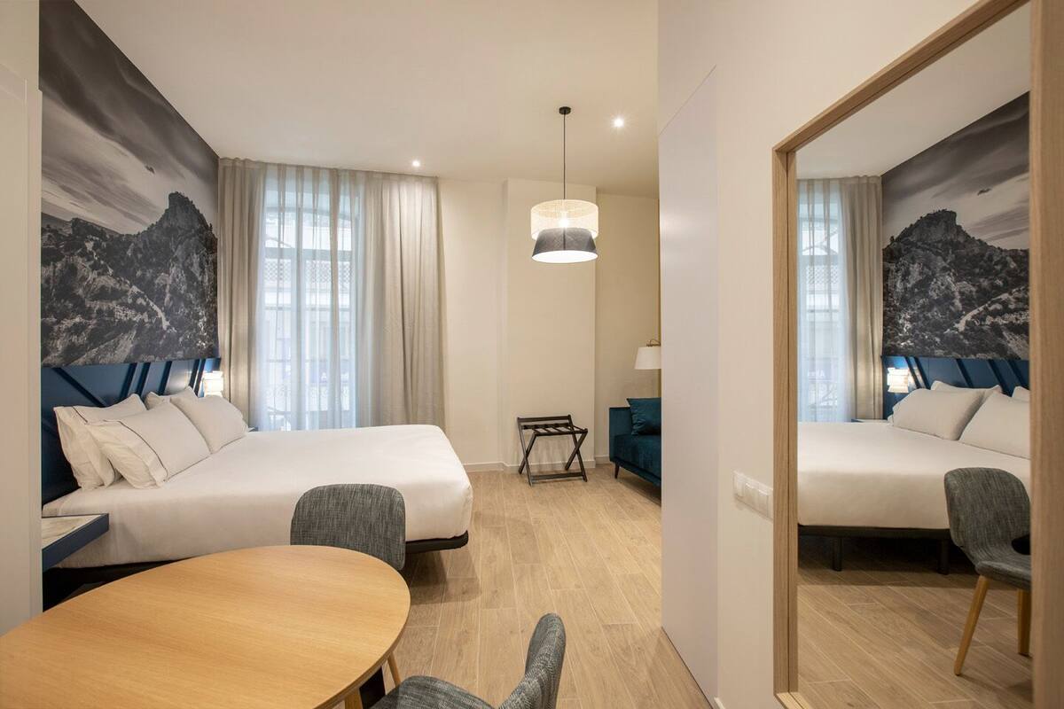 Een hotelkamer met twee bedden, een eettafel en een stoel. Er is een raam met gordijnen, een hanglamp en een wanddecoratie.