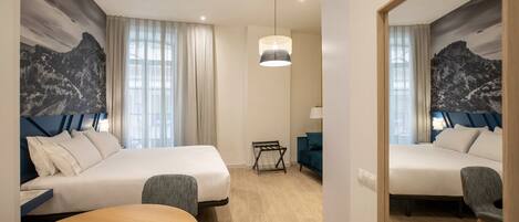 A hotel room with two beds, a dining table, and a chair. There is a window with curtains, a hanging light fixture, and a wall-mounted artwork.