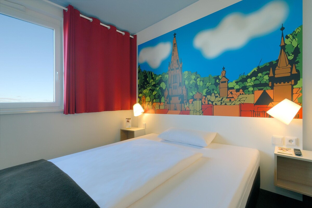 Habitación de hotel con cama, cortinas rojas, un mural de un paisaje urbano y dos lámparas de noche.