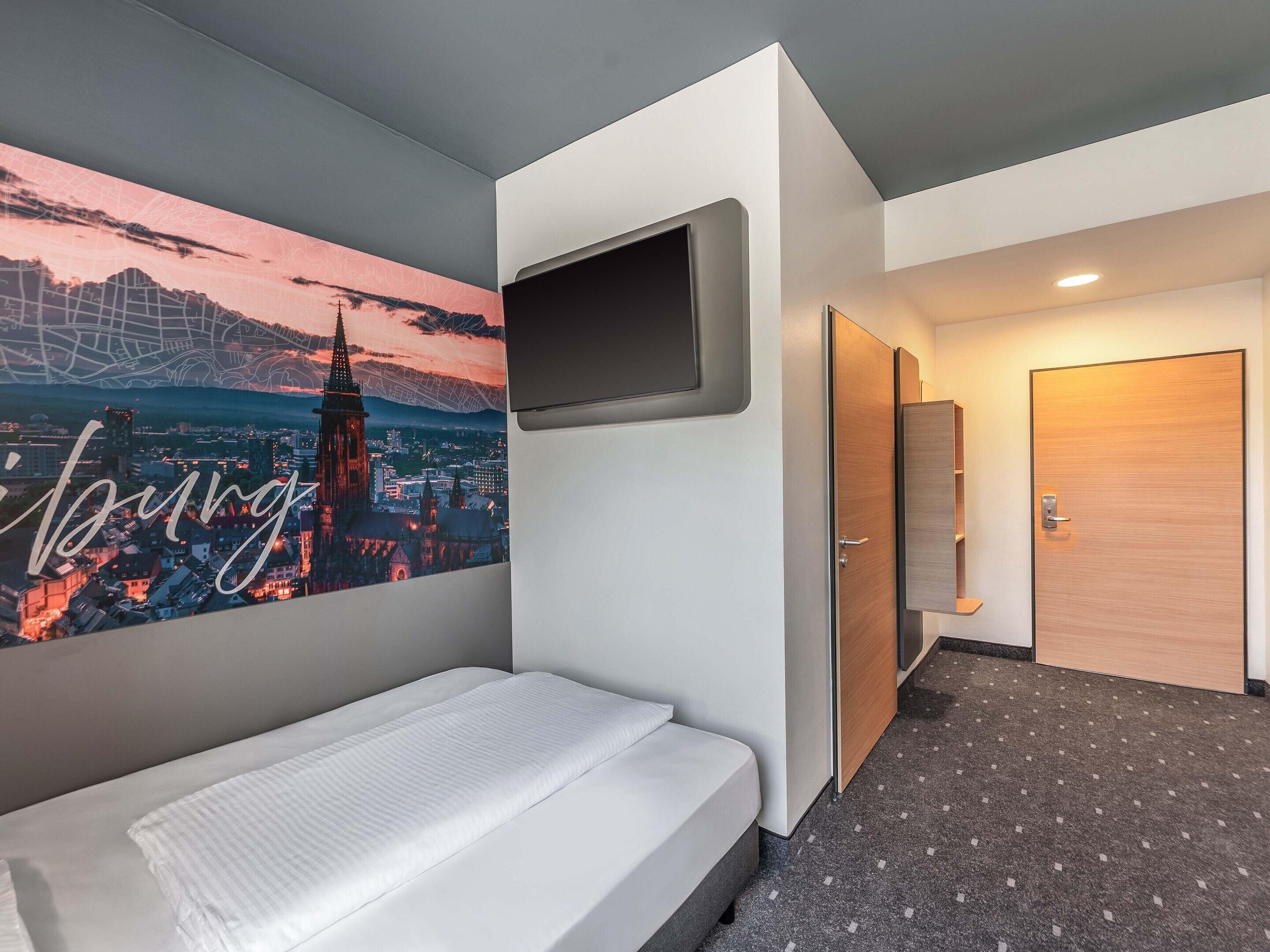 Habitación de hotel con cama, televisor montado, cuadro de paisaje urbano, puerta y piso alfombrado.