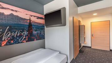 Habitación de hotel con cama, televisor montado, cuadro de paisaje urbano, puerta y piso alfombrado.