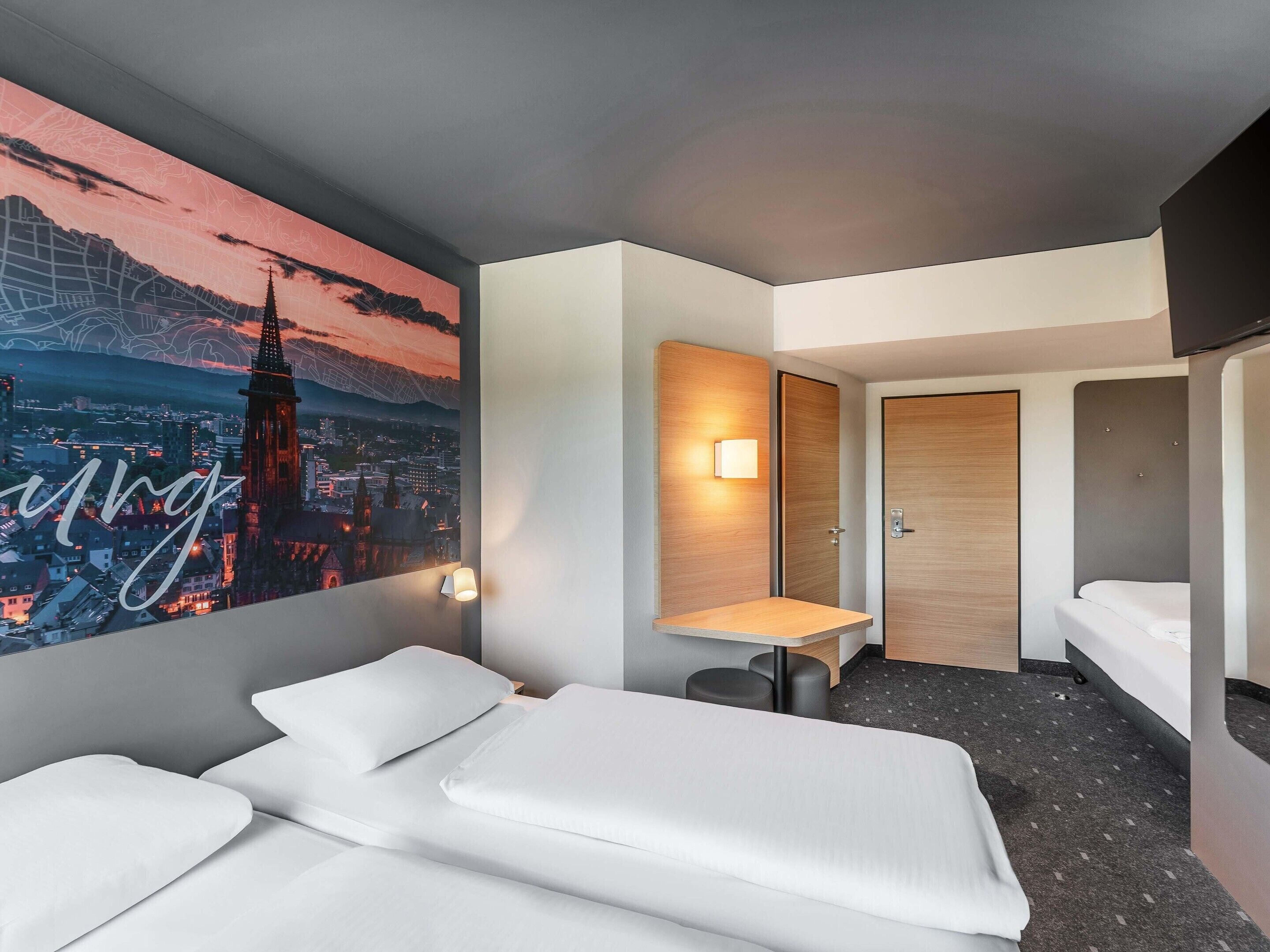 A hotel room with a bed, a desk, a chair, a wall-mounted lamp, and a door.