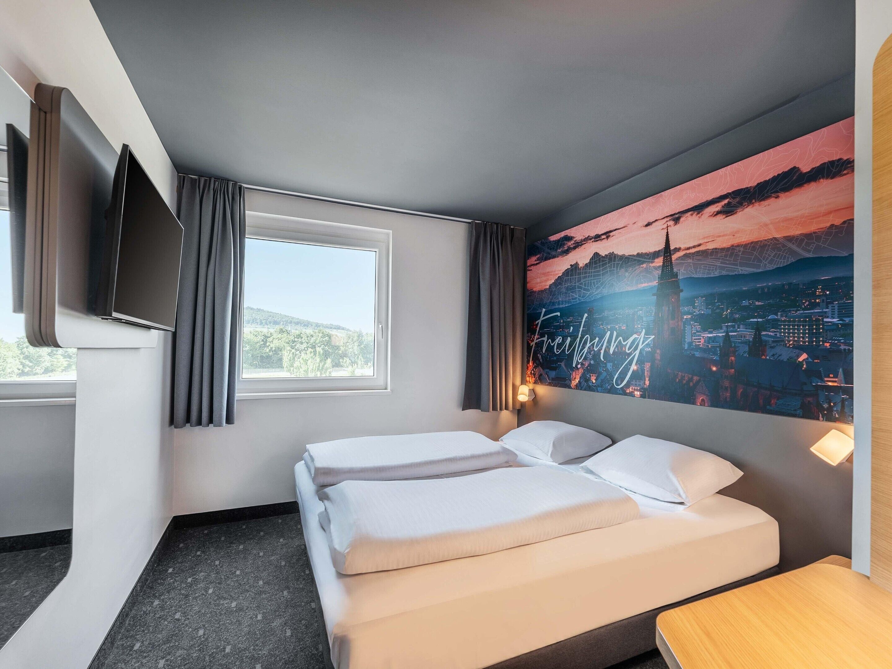 Quarto de hotel com duas camas, uma televisão de tela plana na parede, uma janela grande com cortinas e uma pintura de paisagem urbana na parede.