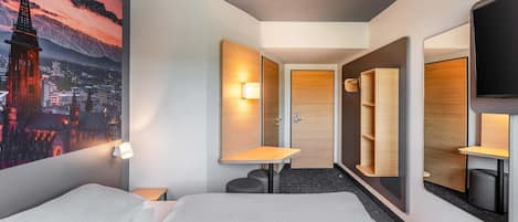 A hotel room with a bed, a desk, a chair, a mirror, and a wall-mounted TV.