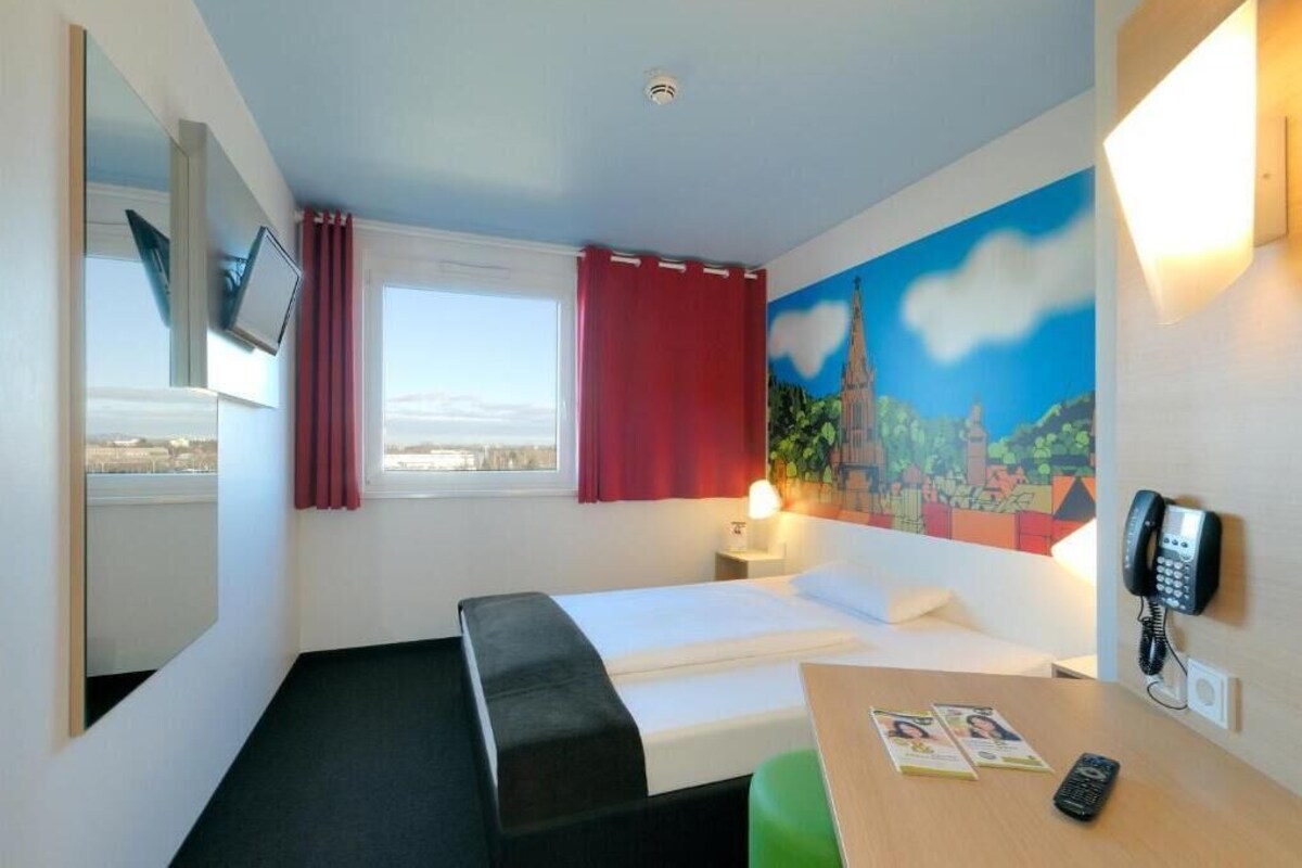 Una habitación de hotel con una cama, un escritorio con teléfono, una ventana con cortinas rojas y un cuadro en la pared.