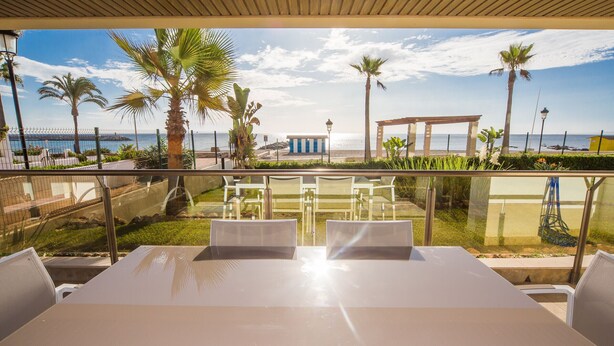 A terrace with a table and chairs, overlooking a beach with palm trees and a clear sky. - Marina del Castillo 305 (Manilva)