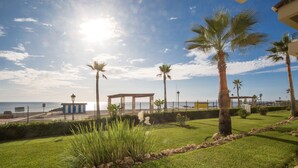 A well-maintained garden with palm trees, a clear view of the sea, and a beach area with a gazebo and a small building. - Marina del Castillo 305 (Manilva)