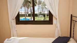 Ein Schlafzimmer mit Bett, einem Fenster mit Vorhängen und Blick auf ein Schwimmbad und Palmen.