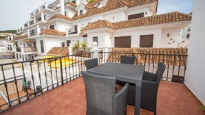 A balcony with a table and chairs, a yellow mat, and a view of white buildings with red-tiled roofs. - Marina Real apto 534 (Manilva)