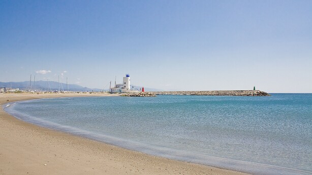 A coastal scene with a lighthouse, clear blue water, and a sandy beach. - Marina del Castillo 302 (Manilva)