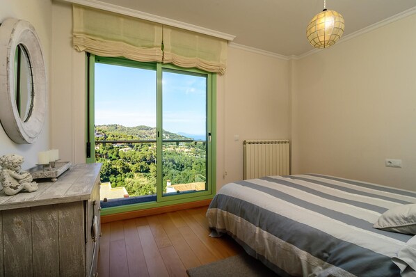 Een slaapkamer met een groot raam, een bed met gestreepte lakens, een kaptafel en een hanglamp.