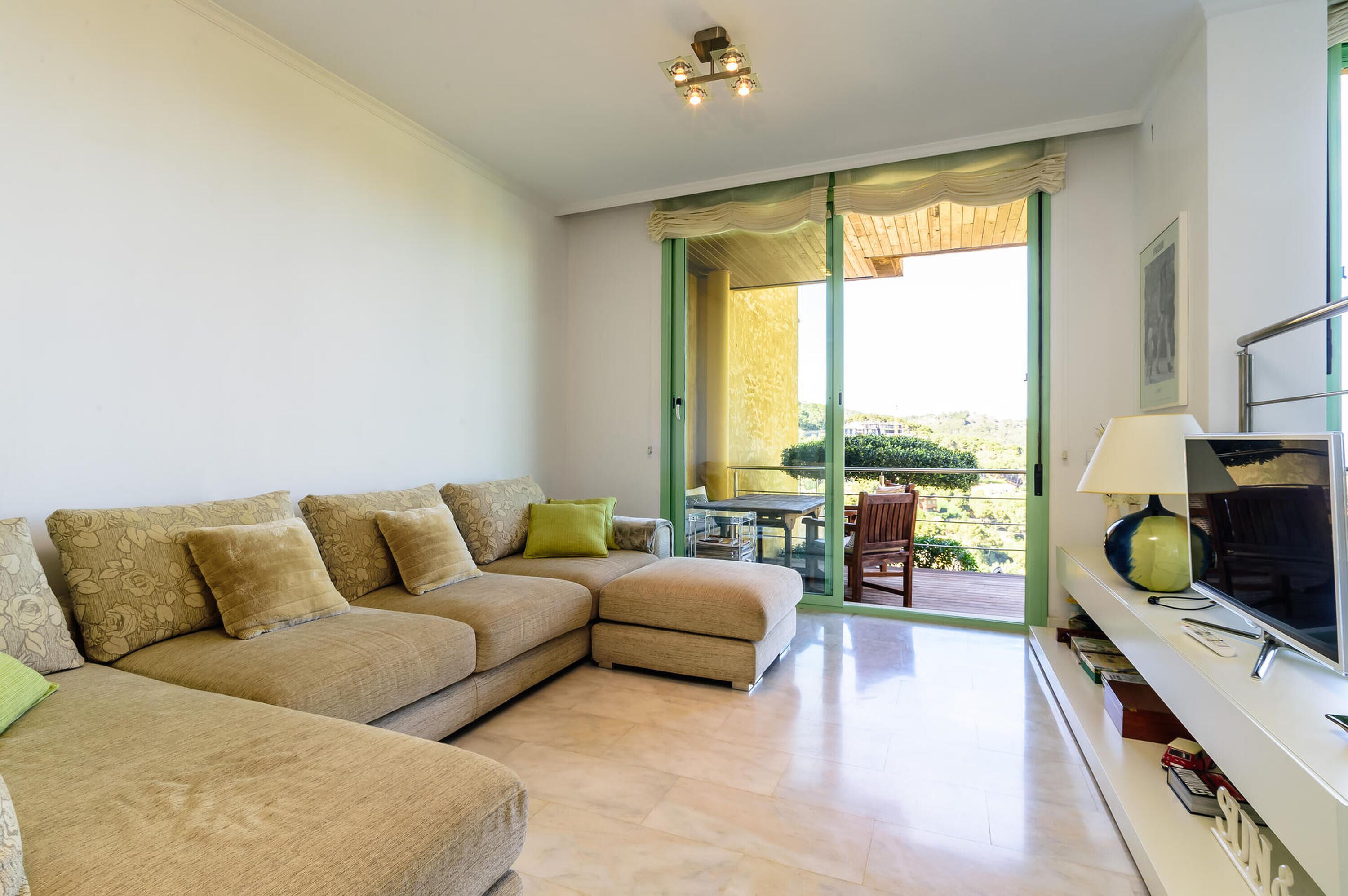 A living room with a beige sectional sofa, a glass door leading to a balcony, and a TV on a white stand.