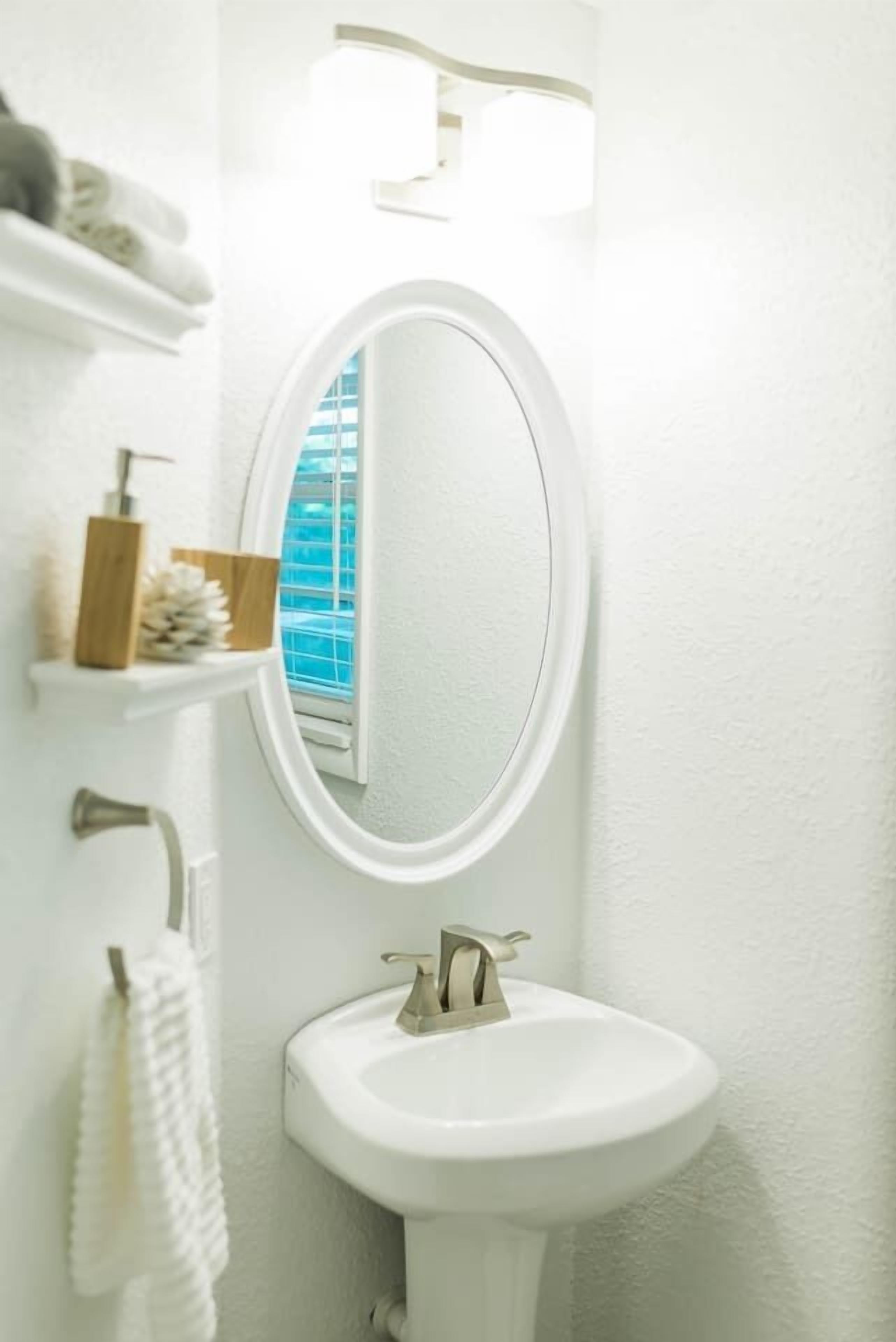 Een badkamer met een wit wastafelmeubel, een ronde spiegel en een raam met blauwe jaloezieën.
