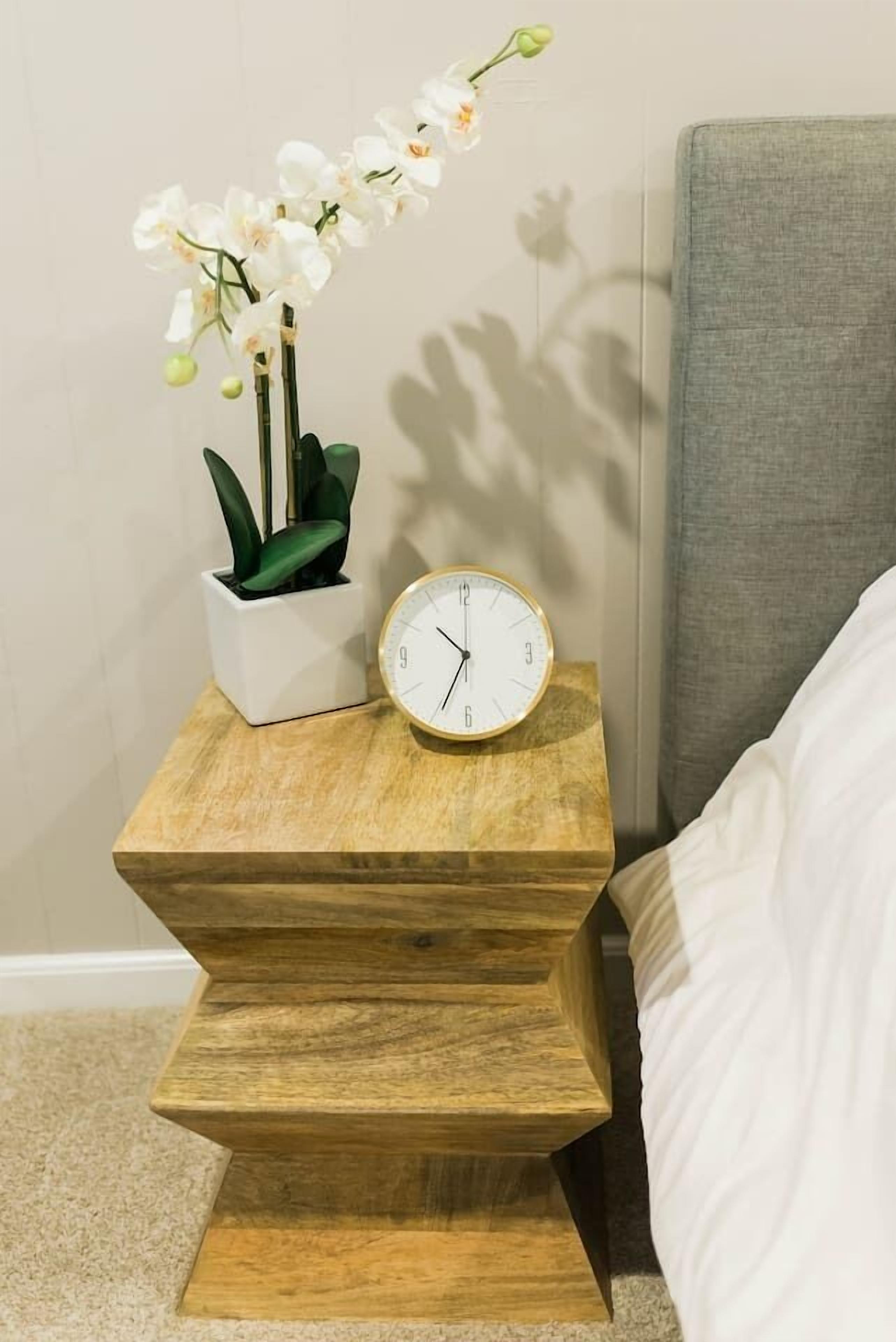 A wooden bedside table with a white clock and a potted orchid.