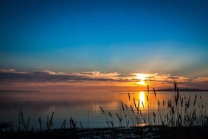 Un coucher de soleil sur une étendue d’eau calme, avec des roseaux au premier plan.