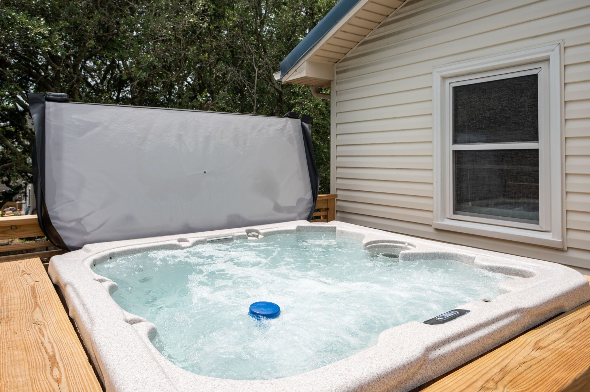 A hot tub with a cover, a wooden deck, and a house with siding and a window.
