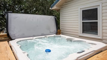 A hot tub with a cover, a wooden deck, and a house with siding and a window.