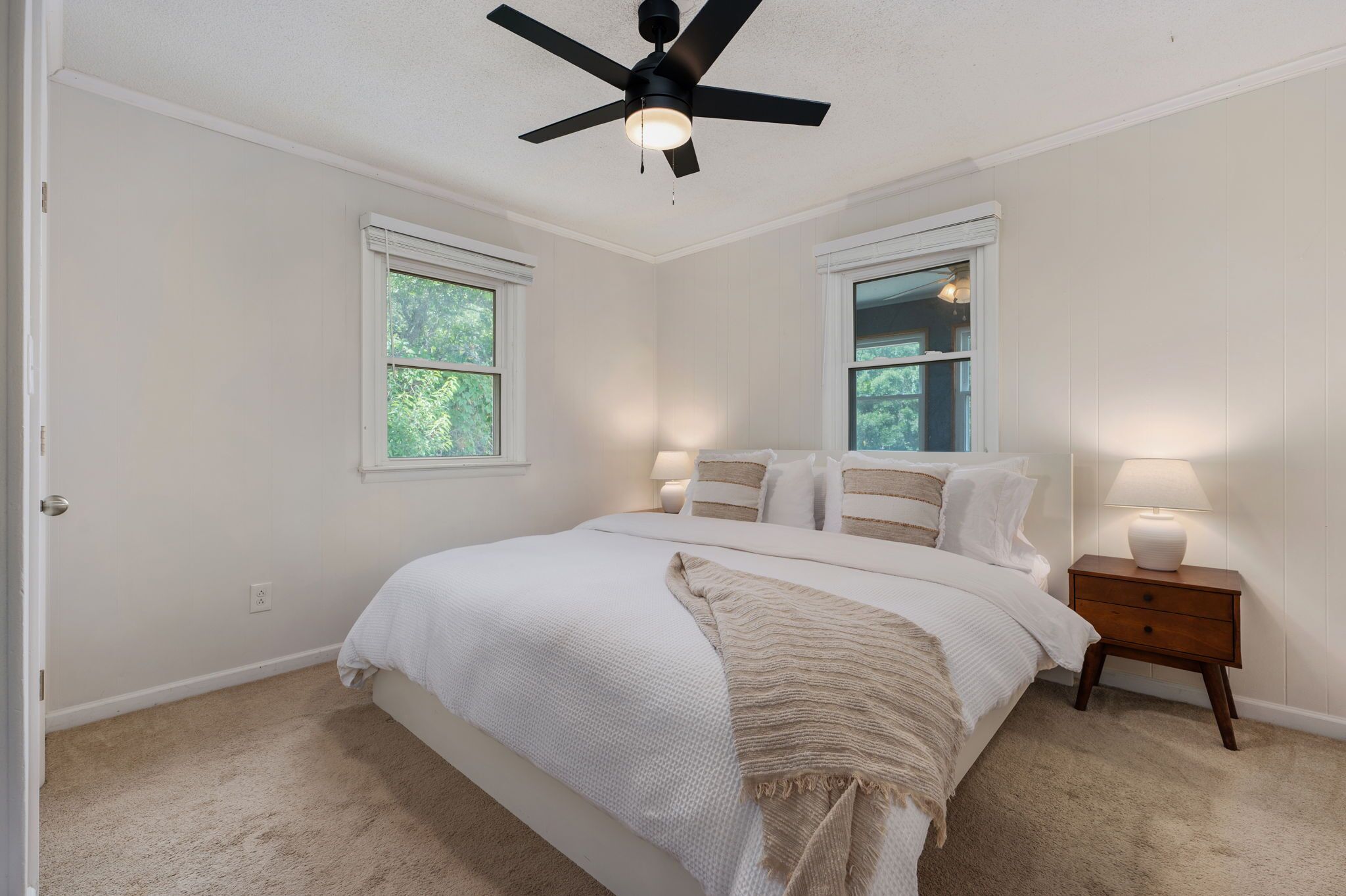 A bedroom with a bed, two bedside lamps, a ceiling fan, and two windows.