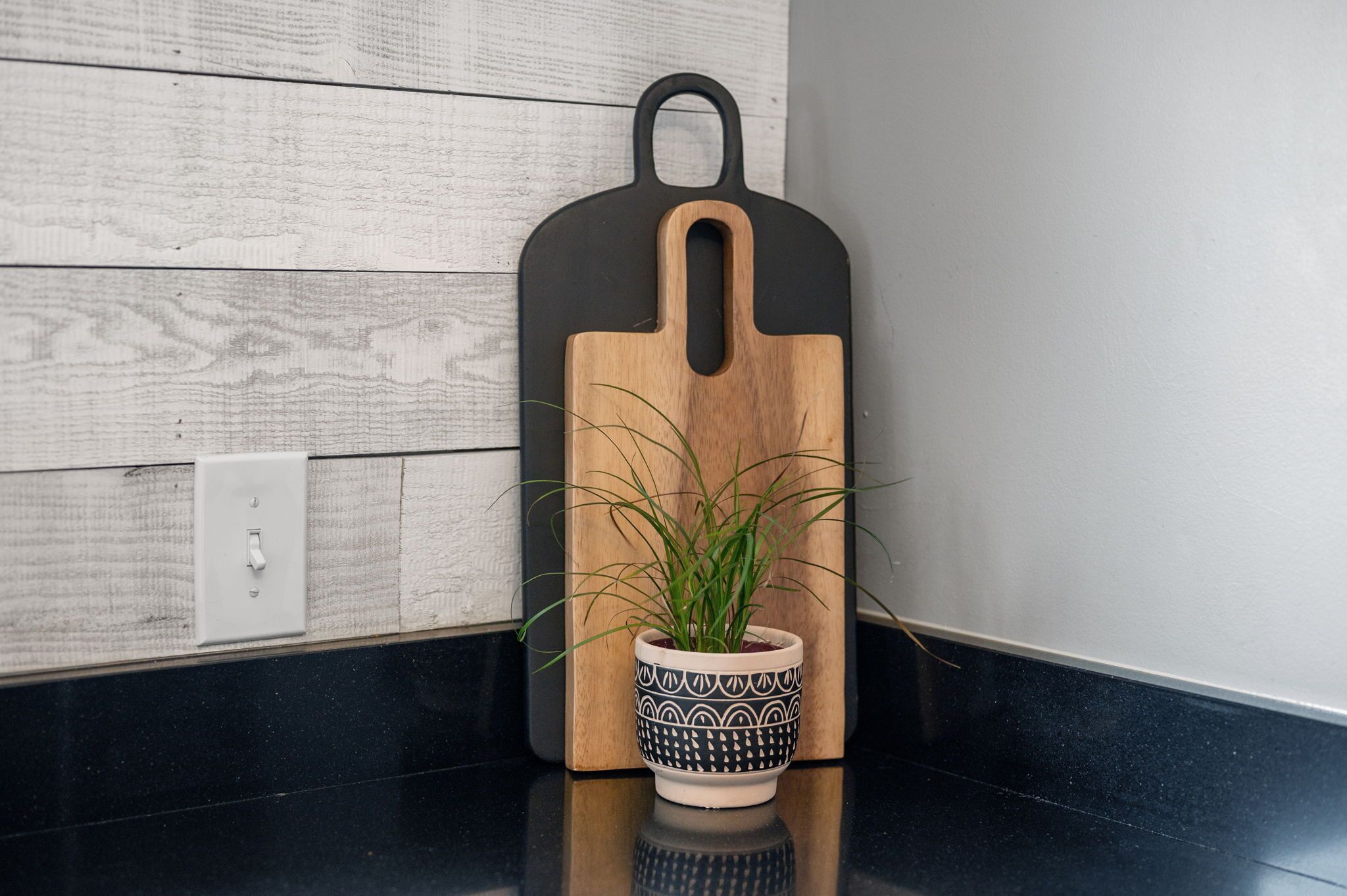 A wooden cutting board with a black handle, a potted plant with long green leaves, and a white switch on a wall.