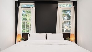 A bedroom with a large bed, two windows with white curtains, and two wall-mounted lamps.