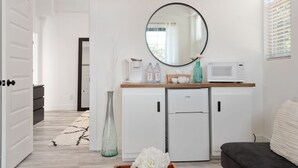 A compact kitchen with a microwave, refrigerator, and white cabinets. A large mirror hangs above the counter, and a vase with flowers is on the floor.