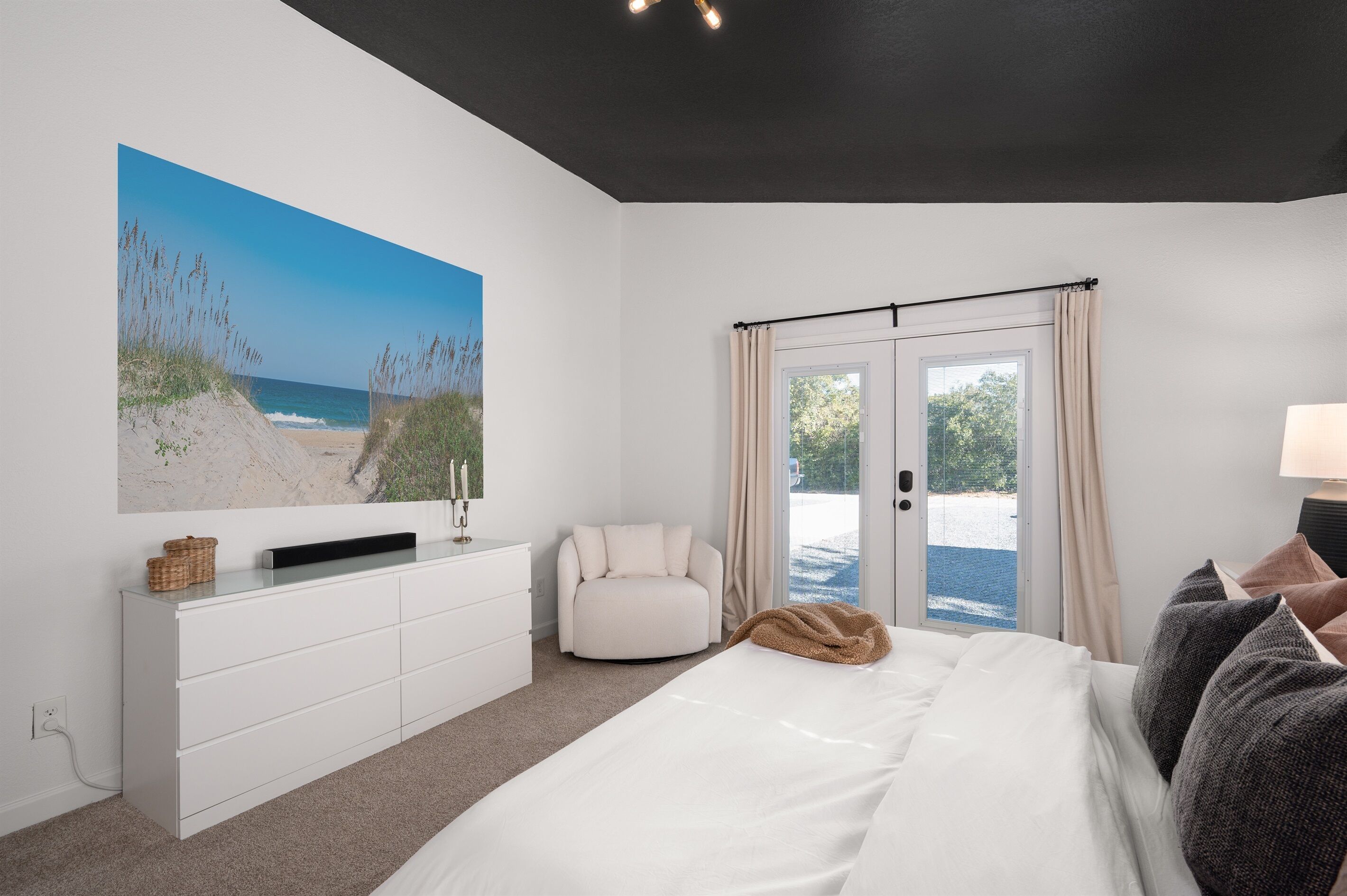 A bedroom with a bed, a white dresser, a white armchair, a large beach painting, and a view of the beach through the door.