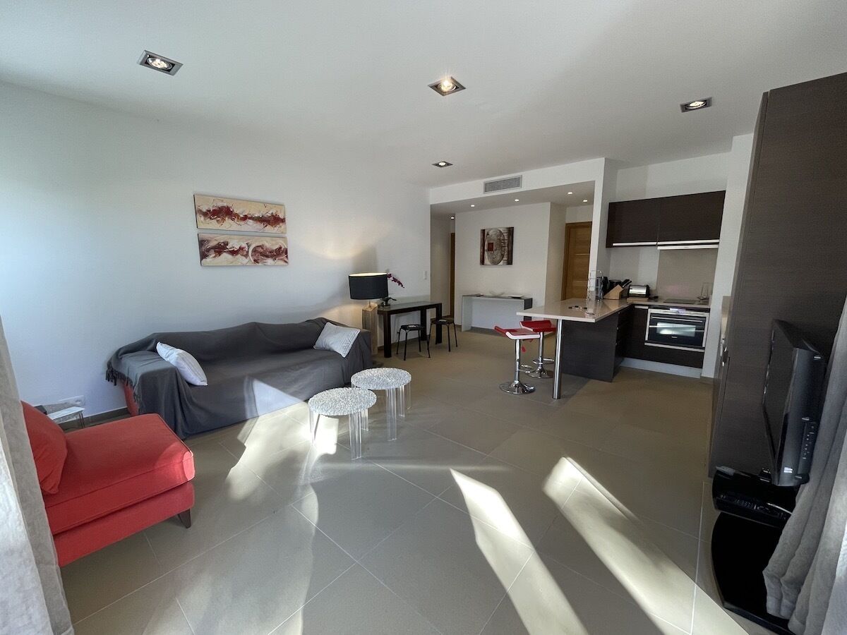 A modern living room with a grey sofa, a red armchair, and a small coffee table. A kitchen area is visible in the background with a dining table and chairs.