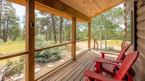 A wooden deck with red Adirondack chairs, large windows, and a view of a forested area. - Hot Tub! Secluded Cabin, overlook Lake Pagosa on 39 acres, No Neighbors! (Pagosa Springs)
