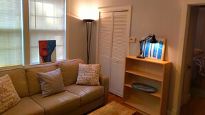 A living room with a beige sofa, a floor lamp, a closed white closet, and a wooden shelf with a blue object. - Urban Retreat on Flint in the Heart of Jonesboro (Jonesboro)