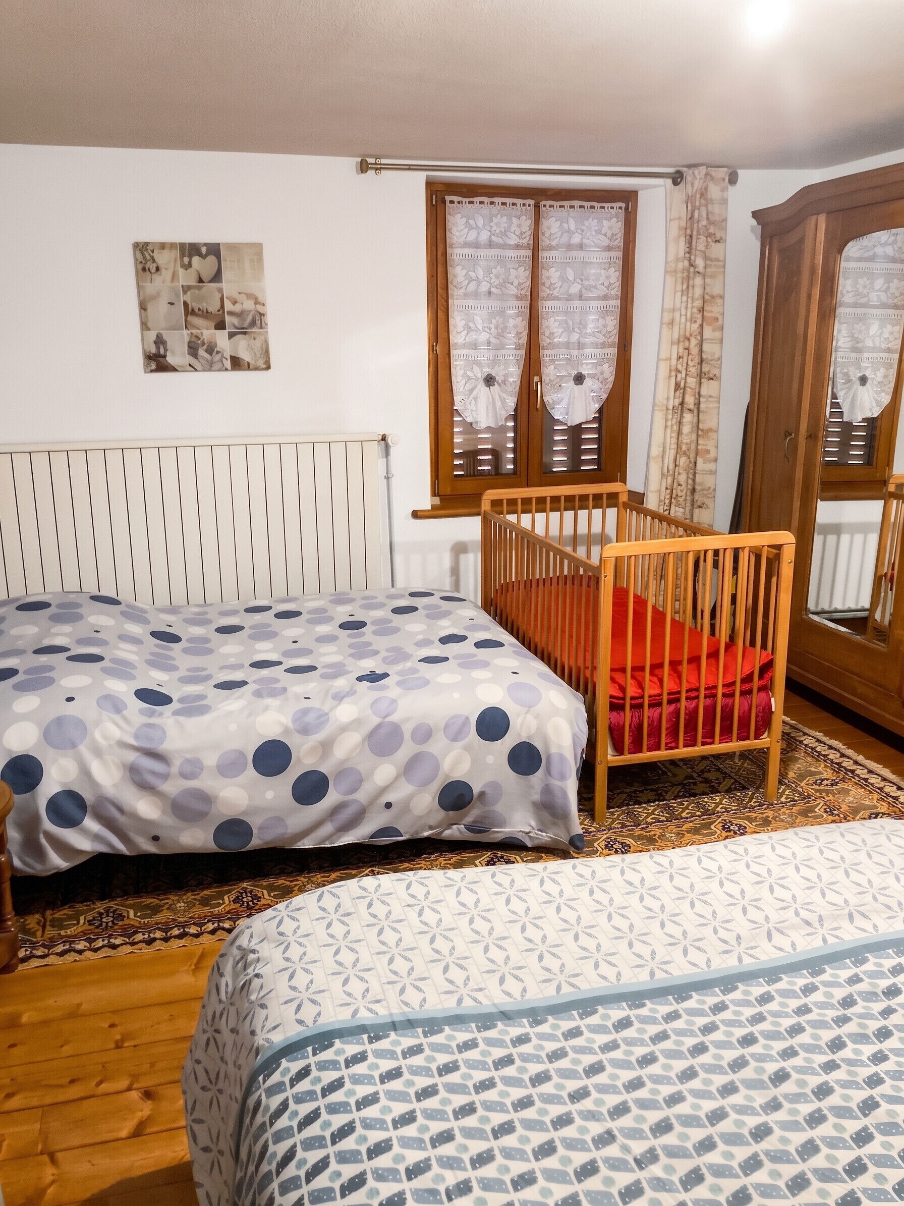 Un dormitorio con dos camas, una cuna de madera, un radiador, una ventana con cortinas y un cuadro colgado en la pared.