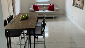Un coin repas avec une table et des chaises, un ventilateur de plafond, une fenêtre avec des stores, un canapé avec des coussins rouges et un tableau au mur.