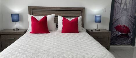 Una cama bien tendida con ropa blanca y almohadas rojas, flanqueada por dos mesitas de noche con lámparas.