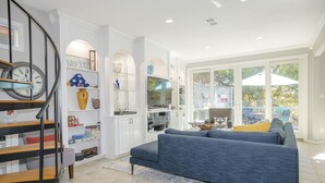 Un salon spacieux avec un canapé bleu, un escalier orné d’une horloge et une grande fenêtre donnant sur un jardin.