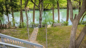 Une terrasse en bois mène à un sentier de pierres qui longe un plan d’eau bordé d’arbres.