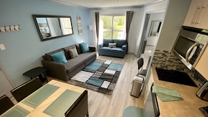 A modern living room with a sofa, a dining table with chairs, a kitchen area with a microwave, and a large window providing natural light. - Lake View Family Condo Disney Universal Orlando (Kissimmee)