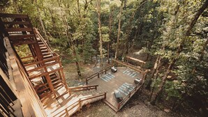 Une terrasse en bois avec un escalier menant à une mezzanine, le tout entouré d’une forêt.
