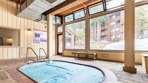 Piscina interior com deck de madeira, placa de aviso e vista de um edifício através de grandes janelas.