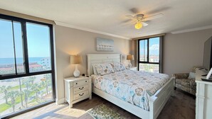 Un dormitorio con una cama grande, dos mesitas de noche, un ventilador de techo y vista al océano.