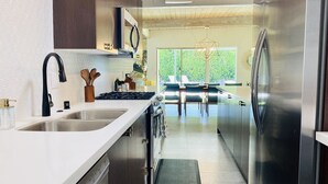 A modern kitchen with stainless steel appliances, a white countertop, and a view of a dining area with a large window. - Paloma House - Mid century style with Frida Kahlo Soul (Palm Springs)
