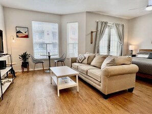 A modern living room with a beige sofa, a white coffee table, and two black chairs. There is a TV on a stand, a lamp, and a plant.