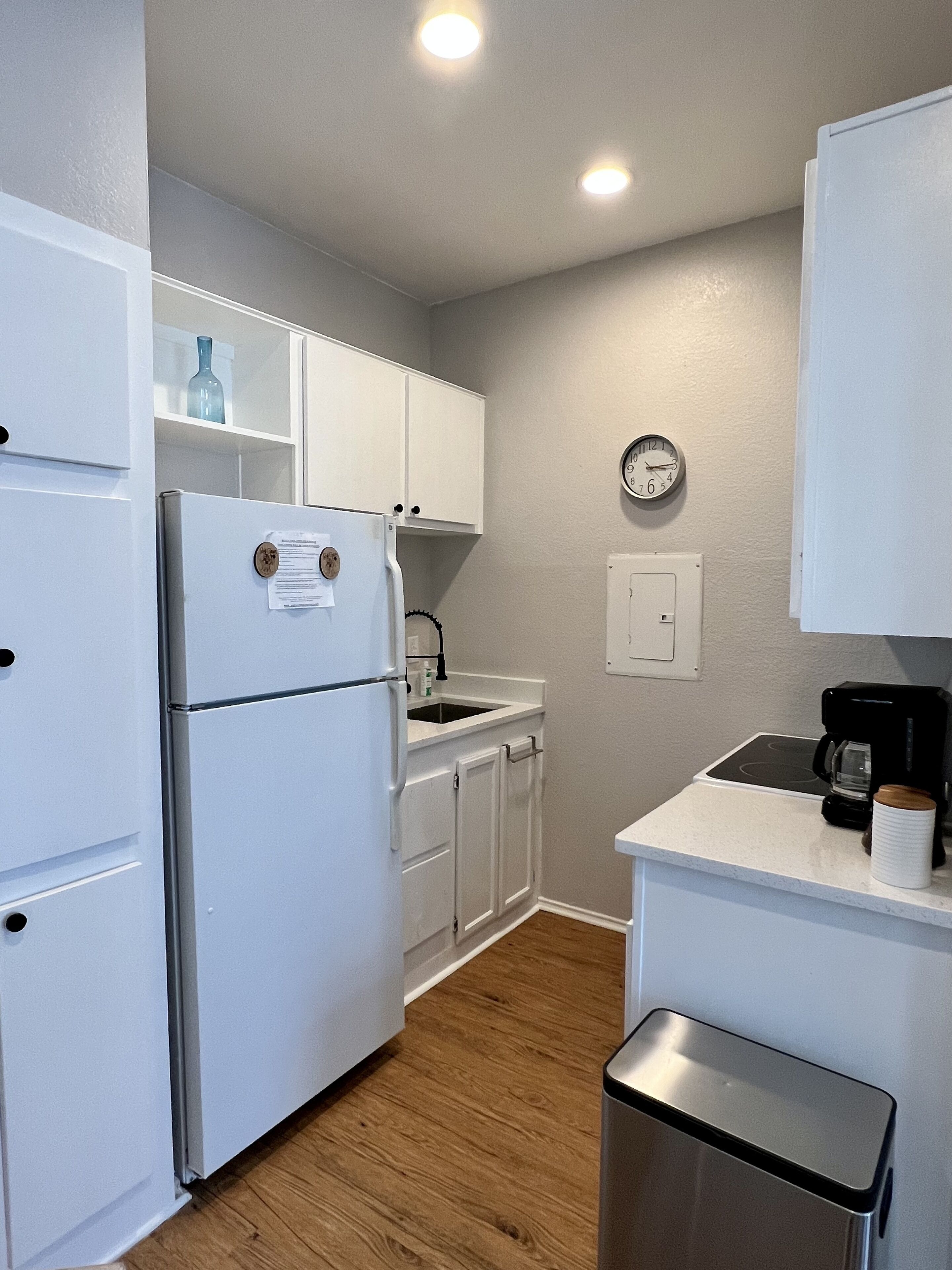 A compact kitchen with white cabinets, a refrigerator, a coffee maker, and a clock on the wall.
